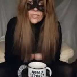 Video of a person with long, straight brown hair, wearing a black mask and black outfit, sitting on a beige couch, holding a white mug with \"Ego Te Robo\" printed on it.