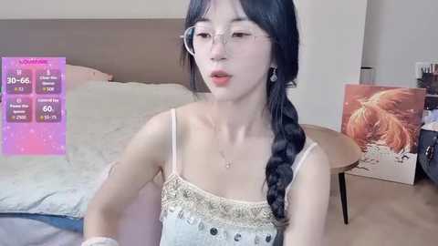 A video of an Asian woman with long black hair in a braid, wearing glasses, a white lace camisole, and a necklace, sitting on a bed in a bedroom with a pink digital display showing exercise metrics.