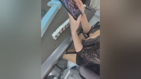 A video of a woman with light skin, wearing black lace lingerie, sitting on a chair, holding a smartphone to take a selfie in a dimly lit room with a mirror.
