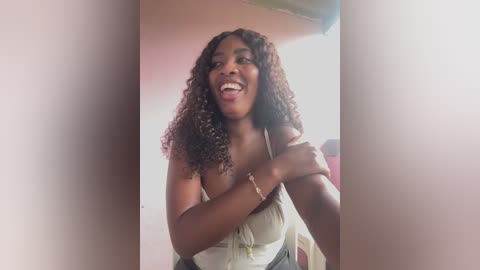 Video of a joyful Black woman with curly hair, wearing a white top and green shorts, sticking out her tongue. She stands indoors with a blurred background.