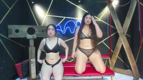 Video of two women in black lingerie kneeling on a red bench, in a dark, geometric-patterned room with neon lights and bondage equipment.