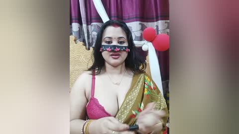Video of a South Asian woman with dark hair, wearing a red bra and a colorful sari, sitting on a wooden chair, with a red blindfold and festive decorations in the background.