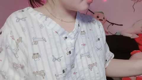 Video of a young person with pale skin and pink hair, wearing a white nightgown with cartoon dog prints. Background features stuffed toys and a pink wall with decals.