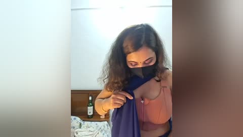 Video of a woman with long brown hair, wearing a pink blouse, and a blue mask, adjusting a blue cloth in a dimly lit room with a wooden nightstand and a bottle of green liquid.