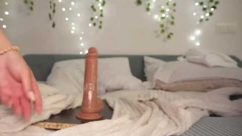 Video of a person's hand with manicured nails holding a large, realistic, flesh-colored dildo on a bed with messy white sheets and pillows, surrounded by soft, glowing fairy lights on a white wall.