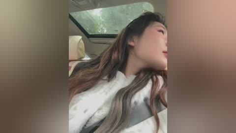 A video of an Asian woman with long, wavy brown hair, wearing a white and black fur-trimmed coat, sleeping in a car seat with a sunroof open, revealing a green tree view outside.