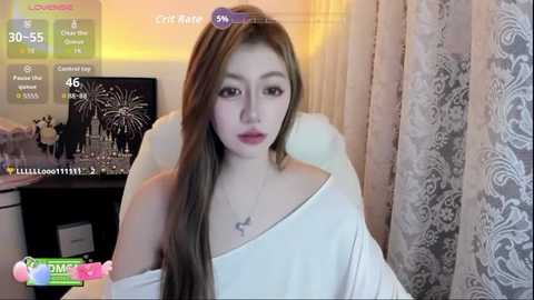 Video of an East Asian woman with long, straight brown hair, wearing a white off-shoulder top, sitting in a room with a dark wooden desk, a computer monitor showing a city skyline, and patterned curtains.