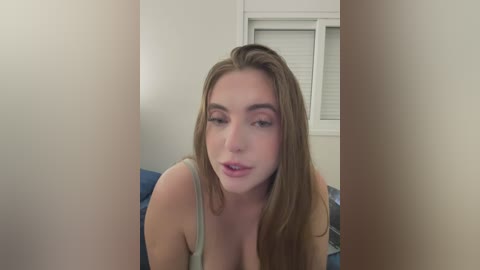A video of a young, fair-skinned woman with long, light brown hair, wearing a green tank top, leaning forward slightly, indoors with white walls and a window with blinds.