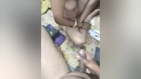 Media: Video of a close-up scene featuring a person's hand holding a penis with a gold bracelet. Background shows a colorful bedspread and a white object.