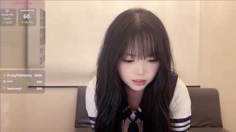 Video of a young Asian woman with long black hair, fair skin, wearing a white shirt and dark tie, sitting on a black leather couch.