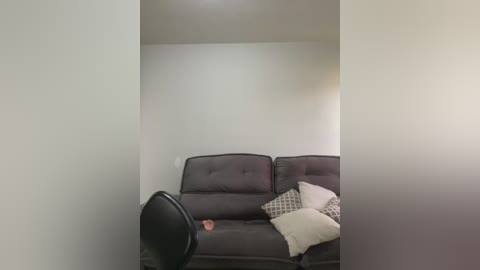 Video of a minimalist, modern living room with a dark gray, tufted sofa, beige throw pillows, and a black swivel chair. The room has white walls and a simple, clean design.