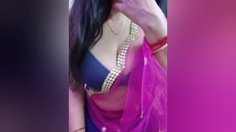 A video of a woman with long black hair, wearing a vibrant pink sari with gold embroidery, and a blue blouse. The background is a gradient of dark to light pink.