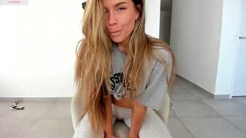 Video of a young, light-skinned woman with long, wavy blonde hair, wearing a grey crop top and white pants, sitting on a white chair in a modern, bright room with white walls and tiled floor.