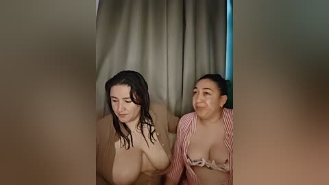 Video of two women with large breasts, one topless, one in a red-striped bra, standing behind a curtain, with blurred background.