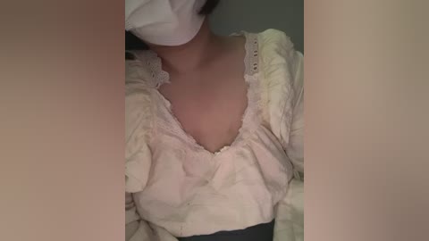 A close-up video of a person wearing a white surgical mask, a white head covering, and a cream-colored, ruffled blouse. The background is blurred and dark, focusing on the upper torso.