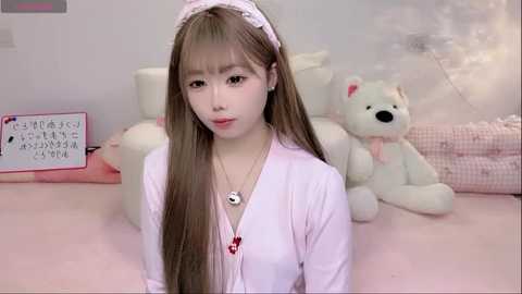 Media: Video of a young Asian woman with long brown hair, wearing a light pink maid outfit, sitting on a bed surrounded by plush toys and a white bear, in a soft, pastel-colored room.