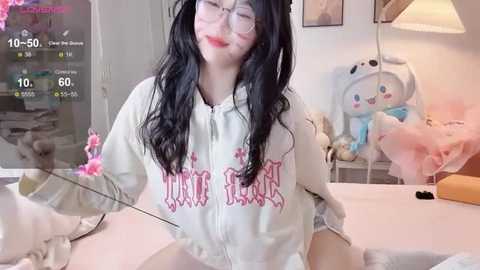 A video of a young Asian woman with long black hair and glasses, wearing a white hoodie with \"MILK\" in red, sitting in a soft-lit, plush-filled room with stuffed toys.