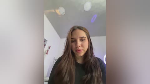 A video of a young woman with long, straight brown hair, wearing a black top, standing in a modern, white-walled room with soft lighting and minimalistic decor.