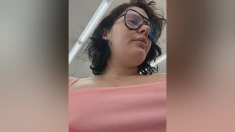 Media: Video of a young woman with short, wavy brown hair, wearing black-framed glasses and a pink off-shoulder top. She has a neutral expression and is captured from a low angle, emphasizing her chest.