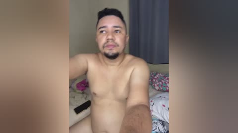 A video shows a nude, muscular, light-skinned man with short dark hair and a beard, sitting on a bed with floral-patterned sheets, a black phone, and a gray curtain in the background.