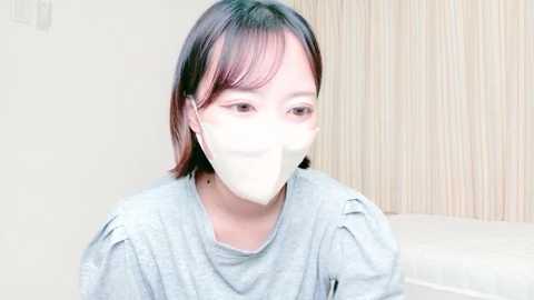 Video of a young Asian woman with shoulder-length dark hair, wearing a light gray long-sleeve shirt and white surgical mask, smiling softly. Background features a beige curtain and a white bed.