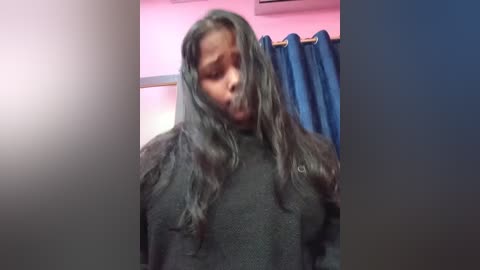 Video of a Black woman with long, wet, dark hair, wearing a black sweater, standing in a bathroom with pink walls and blue curtains.
