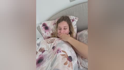 Video of a young woman with light skin and blonde hair, partially covered by a floral-patterned duvet, resting on a light-colored bed with a cushioned headboard.