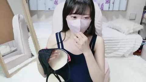 Video of an Asian woman with long black hair, wearing a white mask and dark blue top, applying makeup in a modern bedroom with white walls and a bed.
