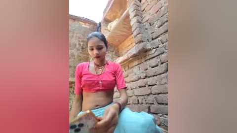 Video of a young woman with medium brown skin, braided hair, wearing a pink crop top and blue skirt, seated in a narrow brick alley, holding a colorful cloth.
