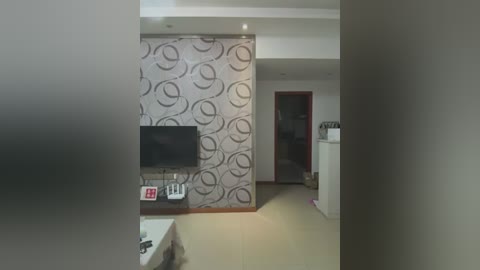 Media: Video of a modern, minimalist living room with a large, abstract-patterned wallpapered wall, a flat-screen TV mounted on it, a small white coffee table, and a white fridge in the background.