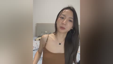 Video of an East Asian woman with long black hair, wearing a brown spaghetti-strap top, large hoop earrings, and a butterfly necklace, standing in a simple bedroom with white walls and a floral-patterned bedspread.