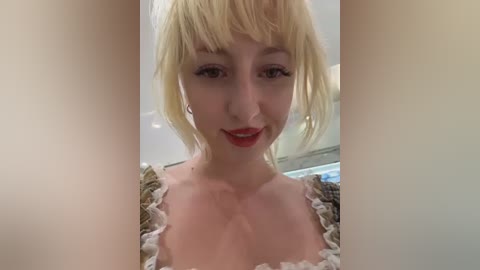 Media: Video of a young woman with platinum blonde hair and bangs, wearing a lace-trimmed dress. She has fair skin, light makeup, and red lipstick. Background shows a blurred indoor setting with soft lighting.