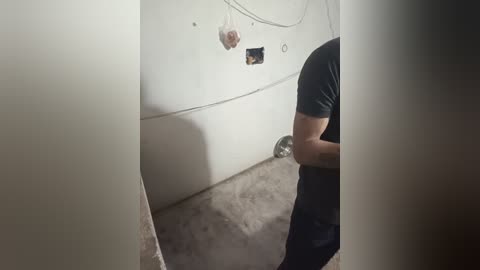 Video of a man in a black shirt, standing in a dimly lit, unfinished room with a white wall, electrical wires, and a metal pole.