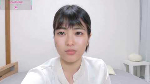 A video of a young Asian woman with short black hair and fair skin, wearing a white shirt, in a minimalist white room with a wooden chair and a white cushion.