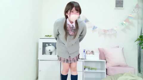 Video of a young woman in a gray school uniform, plaid skirt, and knee-high socks, gagged with tape, standing in a white room with a desk, plants, and decorative flags.