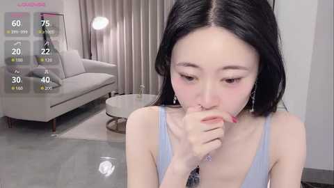 Video of an East Asian woman with long black hair, fair skin, and a slim figure, wearing a light blue tank top, biting her finger in a modern, minimalistic living room with a beige couch and grey floor.