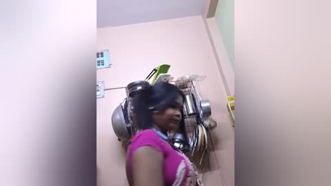Video of a young woman with medium skin tone and straight black hair in a ponytail, wearing a pink shirt and headphones, standing in a narrow, beige-tiled hallway with medical equipment and a green wall.