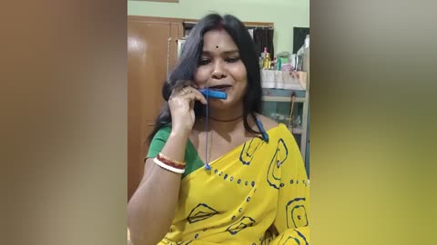Video of an Indian woman with long black hair, wearing a yellow sari with blue patterns, brushing her teeth in a kitchen with wooden cabinets and a countertop cluttered with bottles.