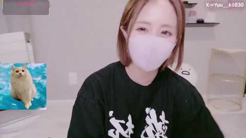 A video of a young woman with light skin and brown hair, wearing a white face mask and a black t-shirt with white characters, standing in a sterile, white room with a cat painting and a transparent chair in the background.