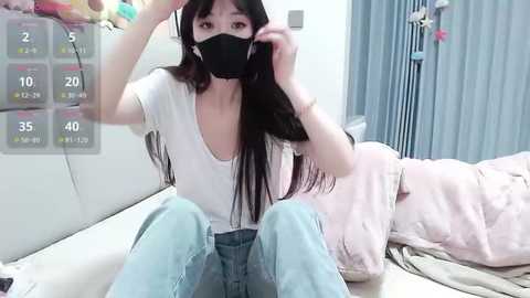 Video of an East Asian woman with long black hair, wearing a black face mask, white top, and blue jeans, sitting on a bed, adjusting her hair.