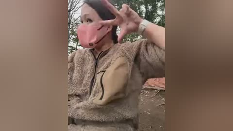 A video shows a woman with a pink face mask, brown fur coat, and brown gloves, making a peace sign. She stands amidst a forest with trees and a red brick building in the background.