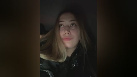 Media: Video of a young woman with light skin and long blonde hair, wearing a black leather jacket, gazing upward with a contemplative expression. The background is dimly lit, creating a moody atmosphere.