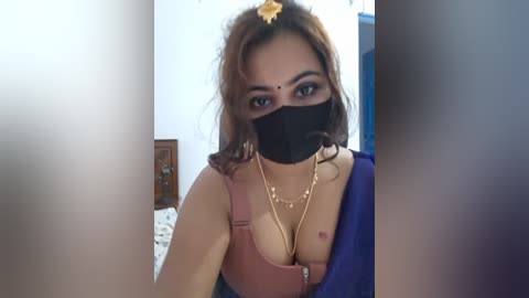Video of a young South Asian woman with light brown skin, wearing a black mask, pink top, and gold necklace. She has a gold bindi and brown hair, with a blue background.