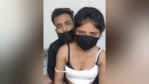 Video of a young couple wearing black masks, with the man in a black shirt and the woman in a white tank top, indoors, blurred background.