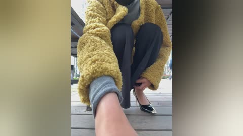 Video of a person in a mustard-yellow fuzzy cardigan, dark pants, and black flats, squatting on wooden stairs. The image is slightly blurred.