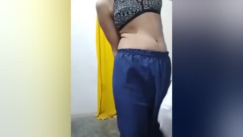 Video of a woman in a black lace bra, blue satin pants, and a yellow garment in a dimly lit room with white walls.