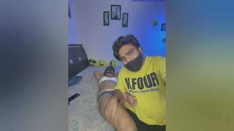 Video of a South Asian man in a yellow shirt, black mask, and glasses, lying on a bed with a woman in a white bra and panties, blurred by a glass partition.