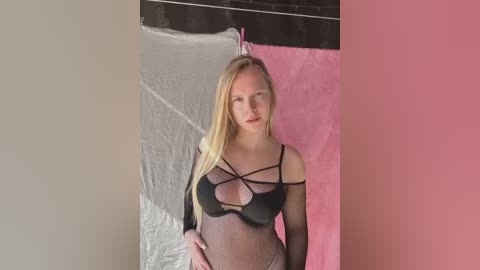 Media: Video of a blonde Caucasian woman with straight hair, wearing black fishnet lingerie with crisscross straps, standing in a studio with a pink and white backdrop.