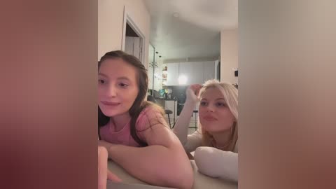 Video of two young women, one with long brown hair, the other with blonde hair, smiling while lying on a bed, partially obscured by a wall. Background shows a dimly lit room with white walls and a kitchen.