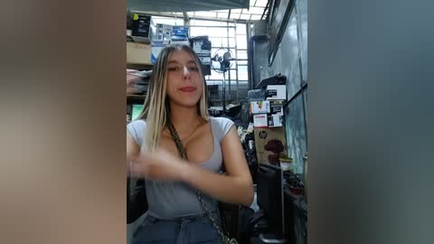 A video of a blonde woman with medium-sized breasts in a low-cut gray top, sitting in a cluttered workshop with shelves of tools and equipment.